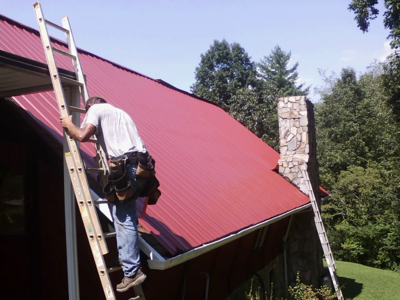 Roofer on a ladder beside a red metal roof and stone chimney — Soffit & Fascia Repair by Roofing Brothers in Dupont